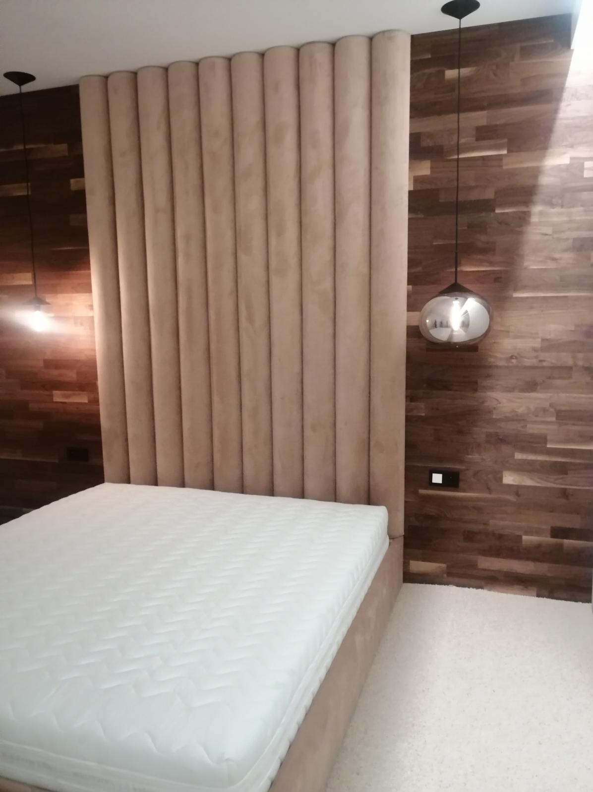 Beżowe, tapicerowane łóżko z wysokim zagłówkiem w pionowe pasy, na tle drewnianej ściany i wiszących lamp.
