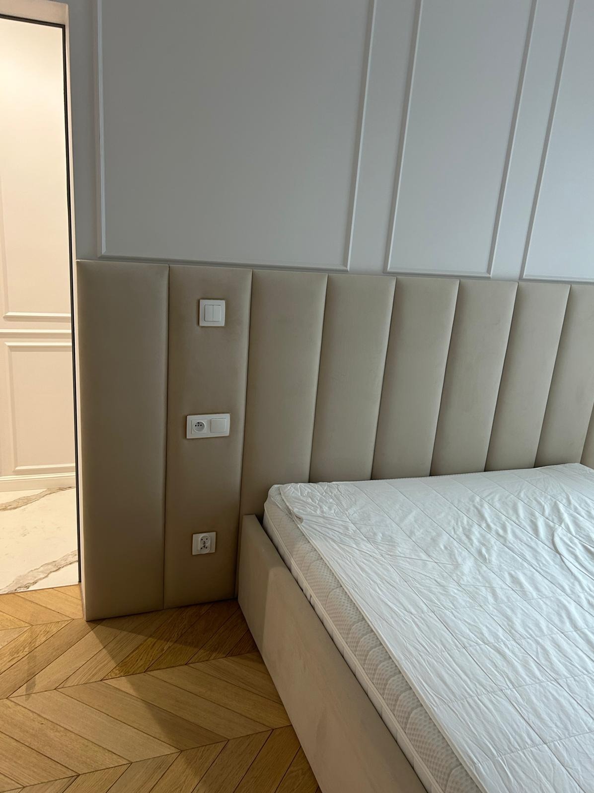 Beżowe, tapicerowane łóżko z wysokim wezgłowiem, panele pionowe. Gniazdka elektryczne na wezgłowiu. Parkiet w jodełkę. Listwy dekoracyjne na ścianie.