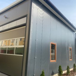 ELEKTRO-TOM - Nowoczesny budynek z szarej blachy trapezowej z automatyczną bramą segmentową i niewielkimi oknami z brązowymi ramami. Widoczne elementy instalacji elektrycznej.