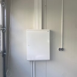 ELEKTRO-TOM - Biała skrzynka elektryczna zamontowana na ścianie z panelami. Widoczne przewody i gniazdka. Ujęcie z wnętrza pomieszczenia.