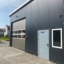 ELEKTRO-TOM - Szara brama segmentowa z oknami w budynku o ciemnej elewacji, obok szare drzwi z tabliczką 'Biuro' i okno. Widok na budynek z zewnątrz, kostka brukowa.