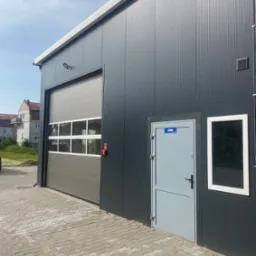 Szara brama segmentowa z oknami w budynku o ciemnej elewacji, obok szare drzwi z tabliczką 'Biuro' i okno. Widok na budynek z zewnątrz, kostka brukowa.