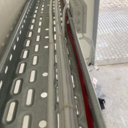 ELEKTRO-TOM - Metalowa korytka kablowa z ułożonymi przewodami elektrycznymi w białej osłonie, zabezpieczonymi plastikowymi opaskami zaciskowymi, oraz czerwonym kablem. Widoczna szara puszka.