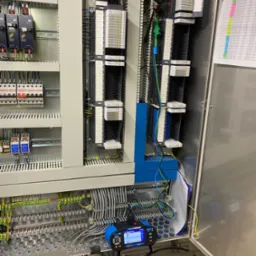 Rozdzielnia elektryczna z modułami, okablowaniem i urządzeniem pomiarowym. Widoczne listwy zaciskowe, przewody w różnych kolorach i dokumentacja.