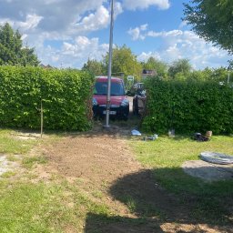 ELEKTRO-TOM - Montaż słupa oświetleniowego na posesji, widoczny czerwony samochód i narzędzia na trawie, żywopłot w tle. Słoneczny dzień.