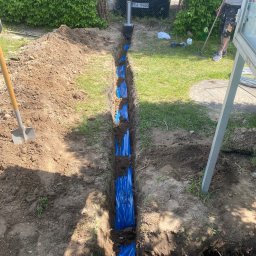 ELEKTRO-TOM - Wykop z niebieskimi rurami w ziemi, szpadel wbity w ziemię obok, trawa, krzewy i samochód w tle. Widoczny etap prac instalacyjnych.