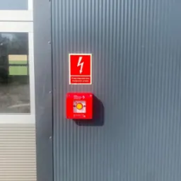 Przeciwpożarowy wyłącznik prądu z czerwoną obudową na szarej ścianie z pionowymi pasami, obok okno z szarą ramą. Oznakowanie bezpieczeństwa.