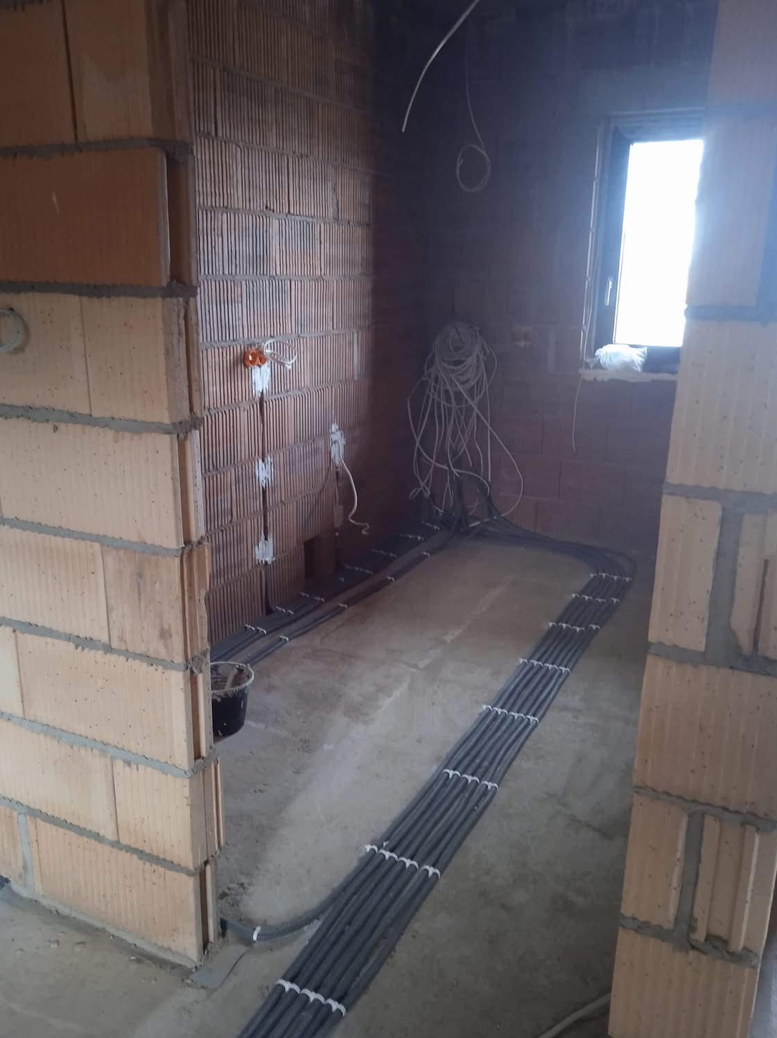 Instalacja elektryczna w rurkach karbowanych, ułożona na podłodze w surowym wnętrzu z ceglanymi ścianami. Widoczne puszki elektryczne i przewody.