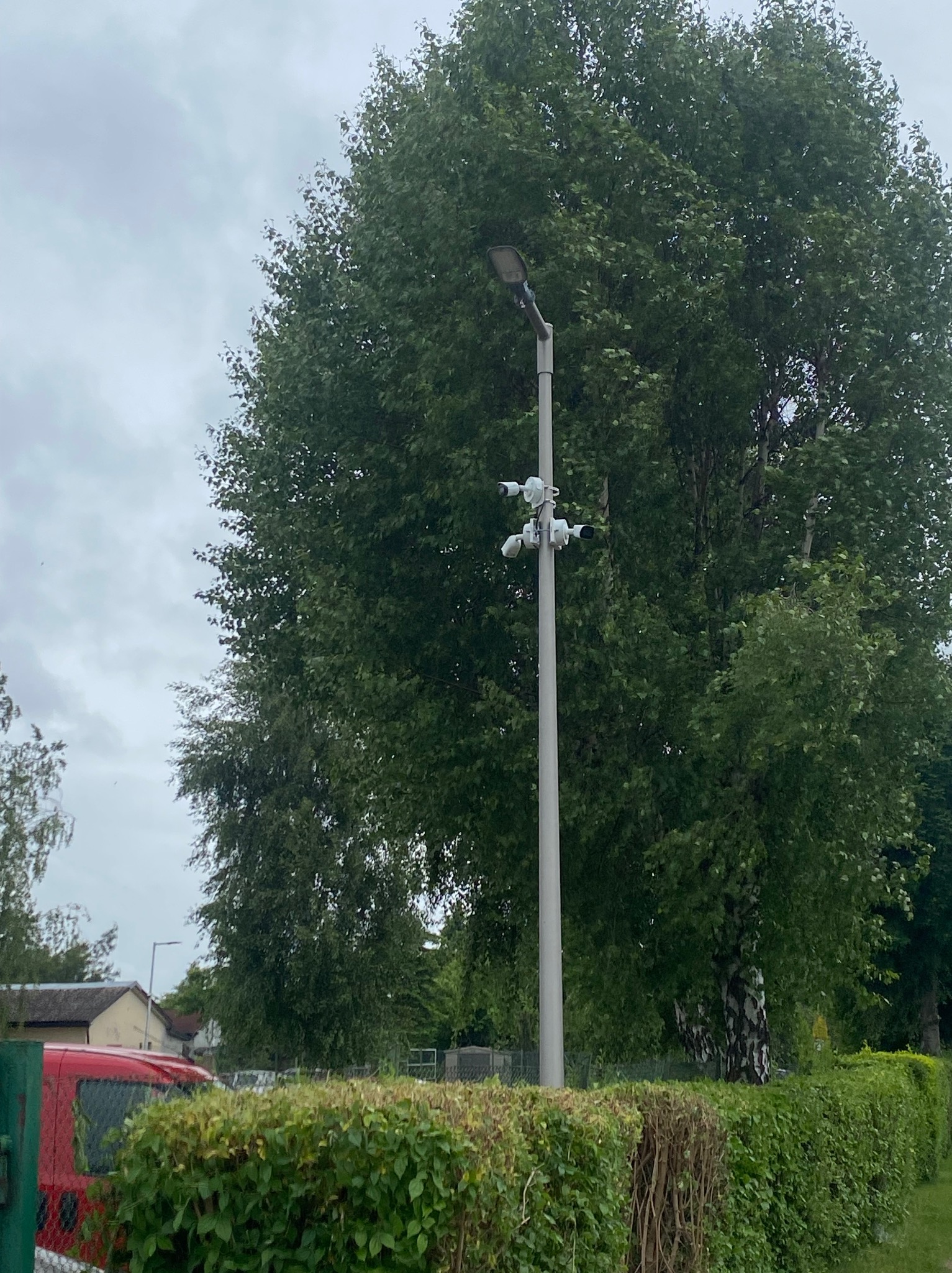 Słup oświetleniowy z zamontowanymi kamerami monitoringu na tle zieleni i pochmurnego nieba, widoczny fragment czerwonego samochodu.