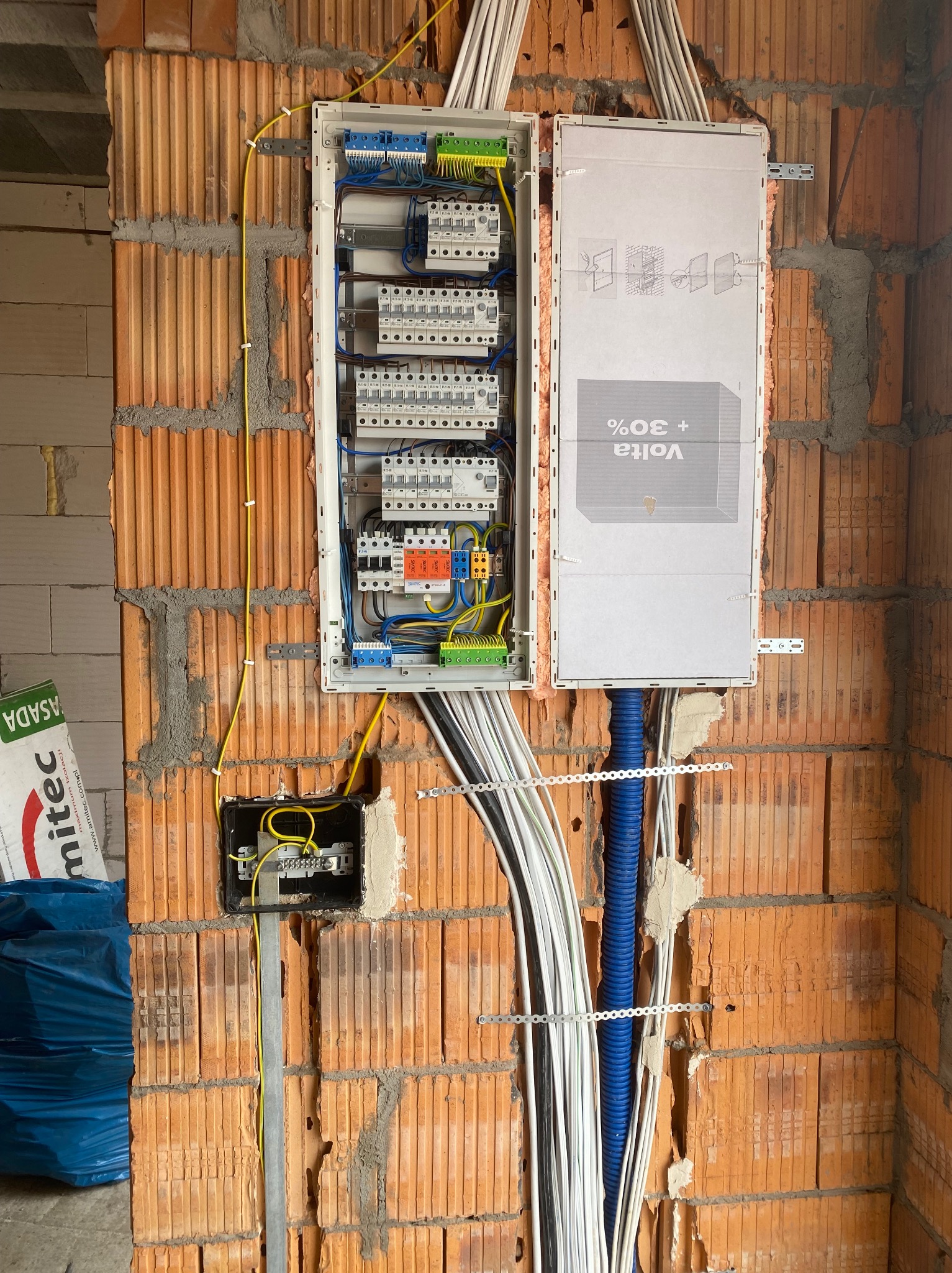 Otwarta skrzynka elektryczna z bezpiecznikami i przewodami na ceglanej ścianie w trakcie instalacji. Widoczne kable w kolorach: biały, żółty, niebieski i czarny.