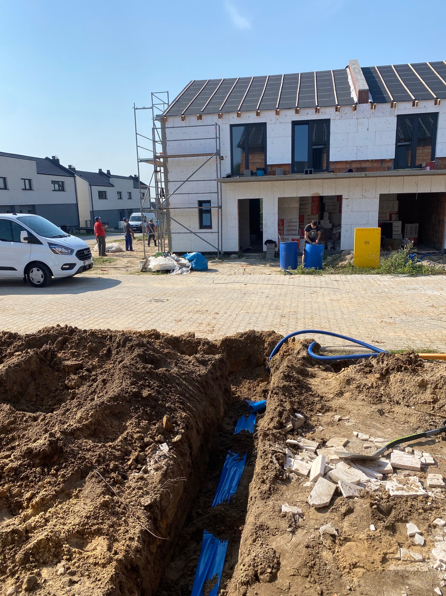 Wykop z niebieskimi rurami w ziemi na tle budowy domu w zabudowie szeregowej. Widoczni pracownicy i samochód dostawczy.