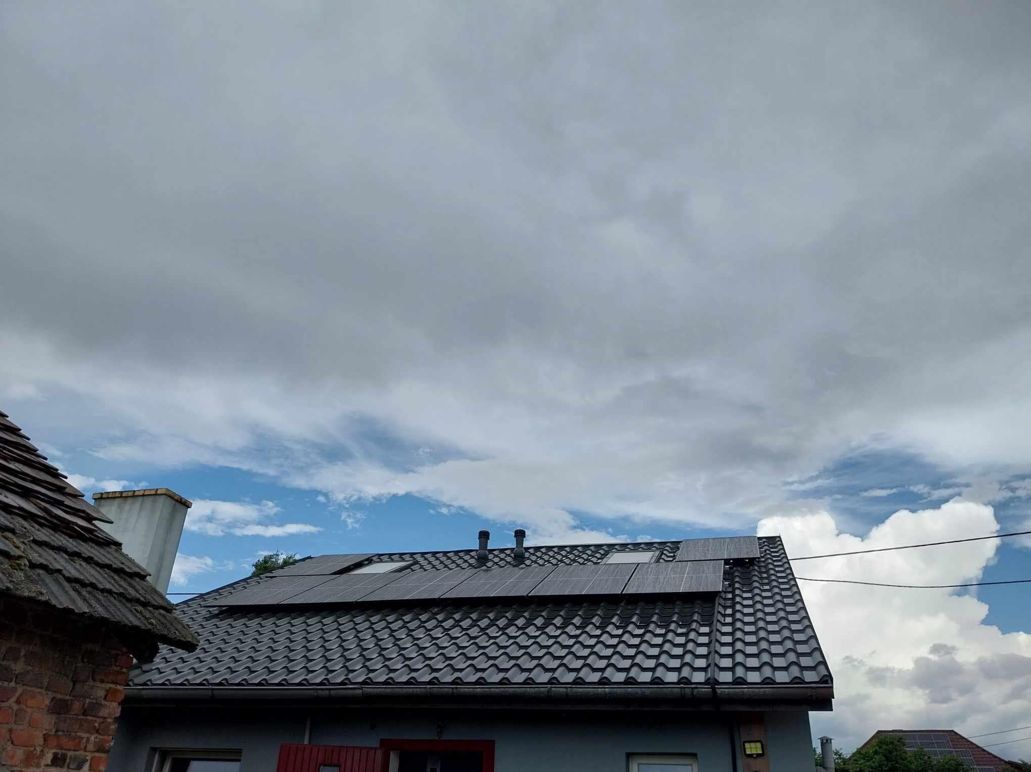 Czarny dach z panelami słonecznymi w pochmurny dzień. Widoczne okna dachowe i kominy. Częściowo widoczny budynek z szarej cegły i błękitne niebo z chmurami.