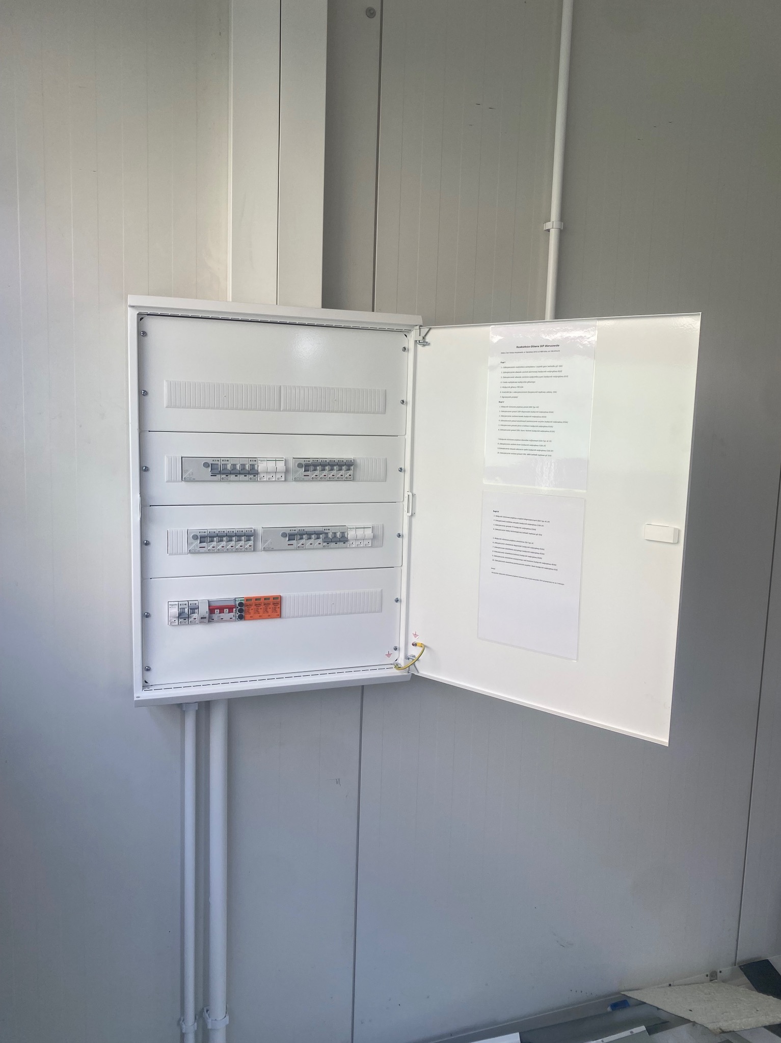 Otwarta skrzynka elektryczna z wyłącznikami i dokumentacją techniczną na białej ścianie z panelami. Widoczne przewody i rury instalacyjne.