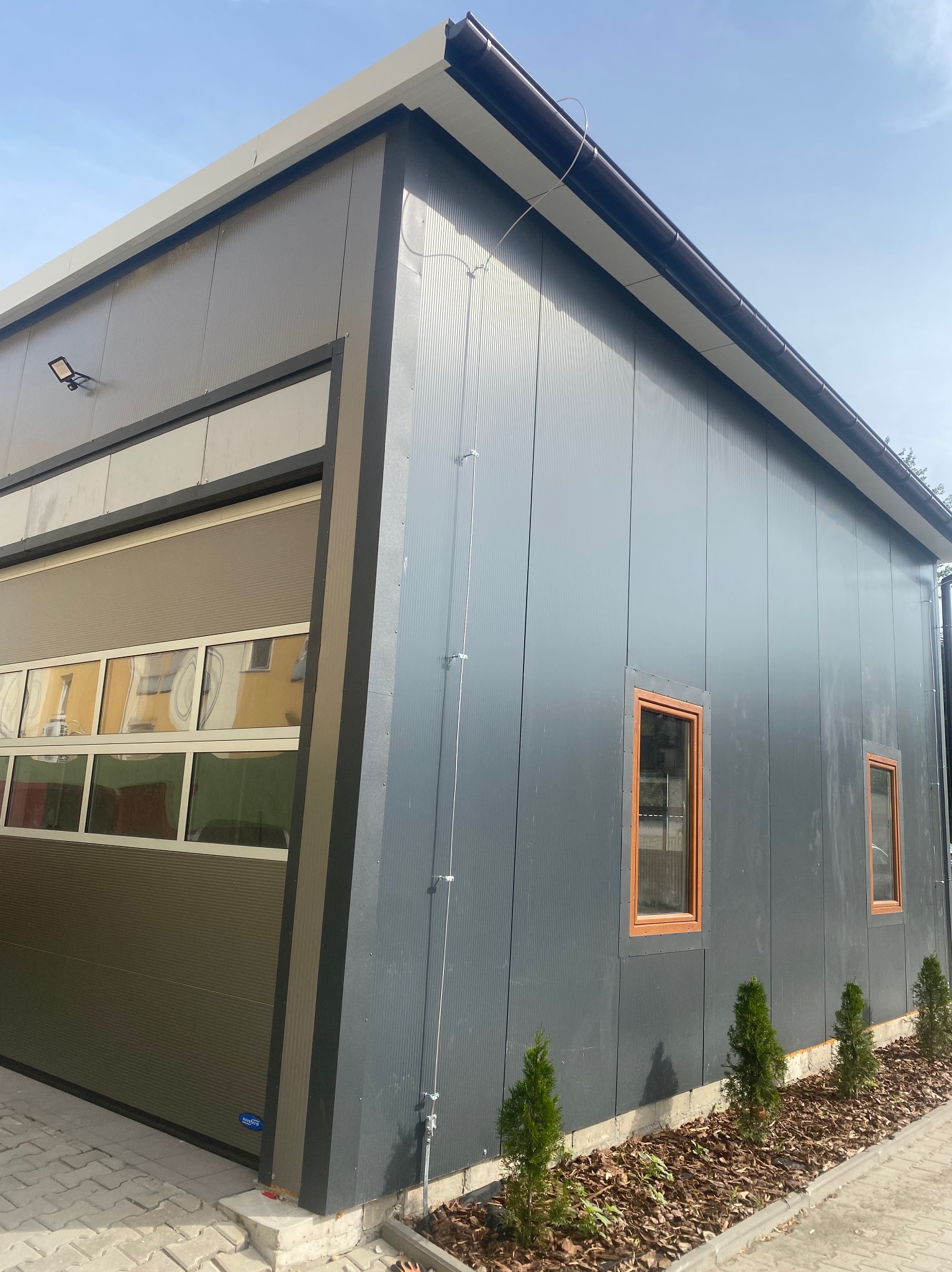 Nowoczesny budynek z szarej blachy trapezowej z automatyczną bramą segmentową i niewielkimi oknami z brązowymi ramami. Widoczne elementy instalacji elektrycznej.