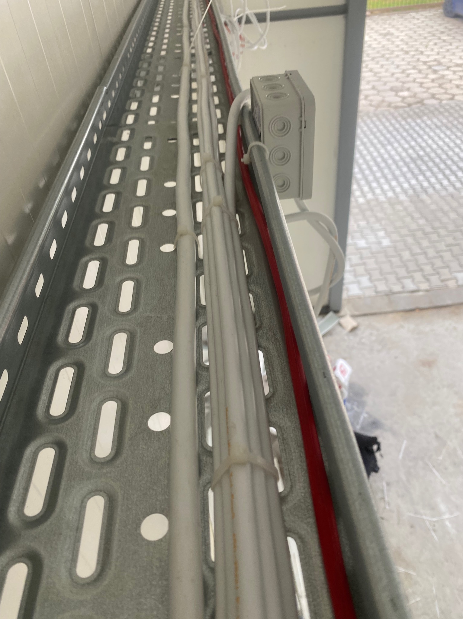 Metalowa korytka kablowa z ułożonymi przewodami elektrycznymi w białej osłonie, zabezpieczonymi plastikowymi opaskami zaciskowymi, oraz czerwonym kablem. Widoczna szara puszka.
