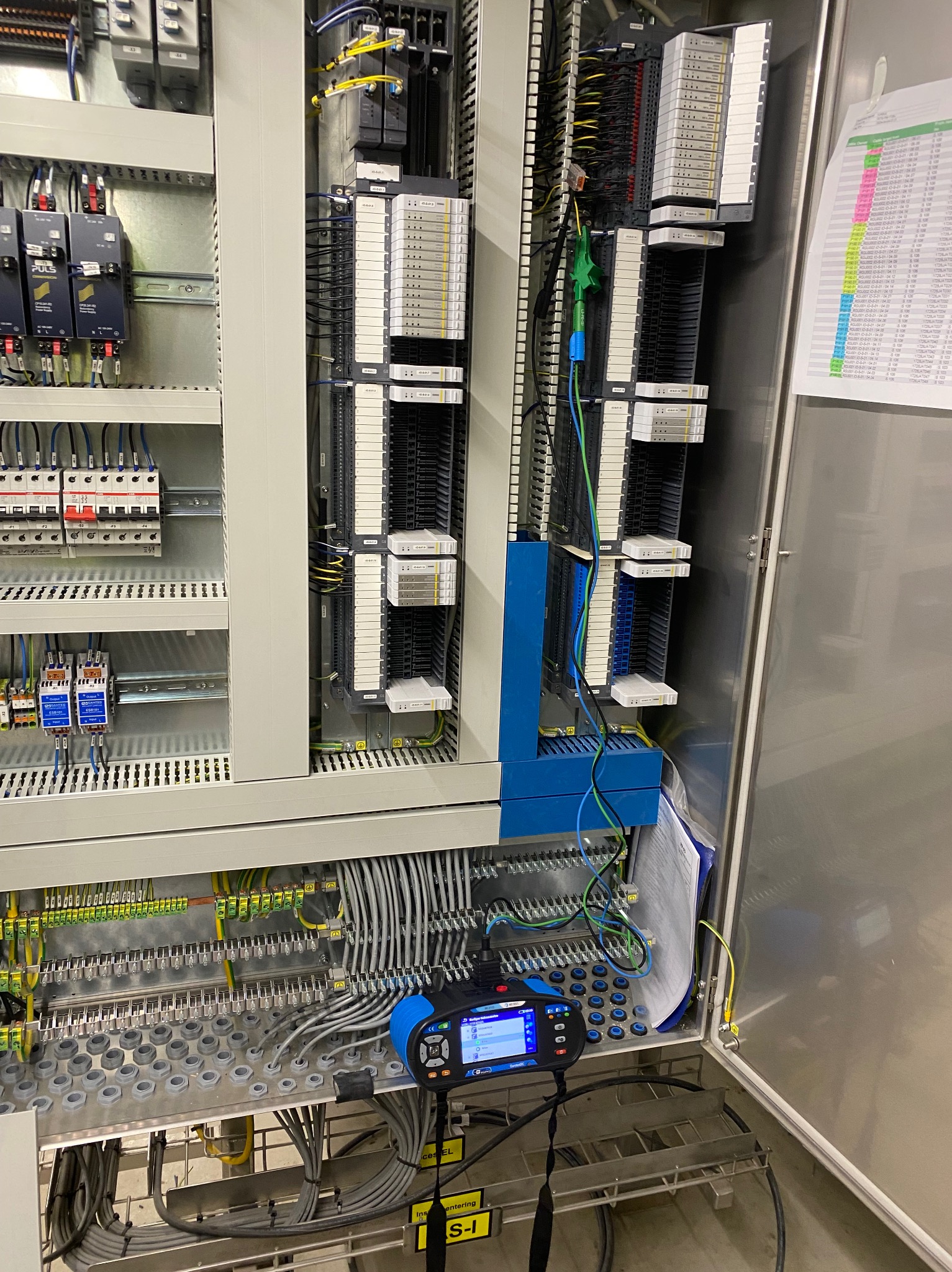 Rozdzielnia elektryczna z modułami, okablowaniem i urządzeniem pomiarowym. Widoczne listwy zaciskowe, przewody w różnych kolorach i dokumentacja.