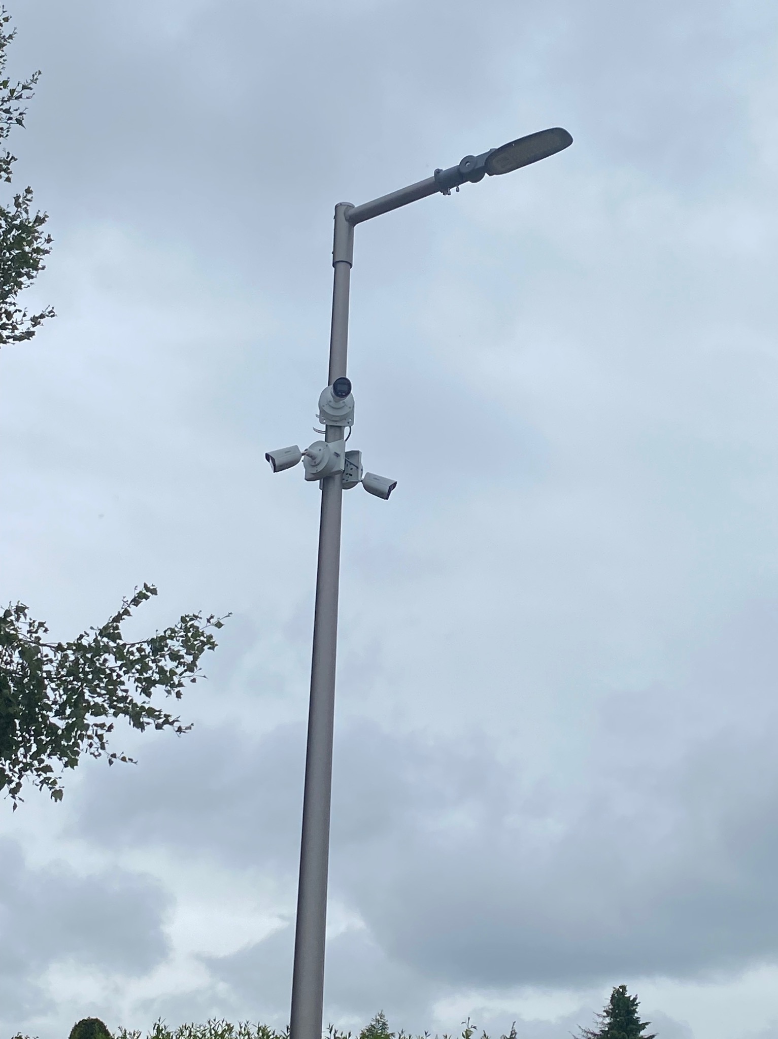 Słup oświetleniowy z zamontowanymi trzema kamerami monitoringu skierowanymi w różne strony, na tle pochmurnego nieba.