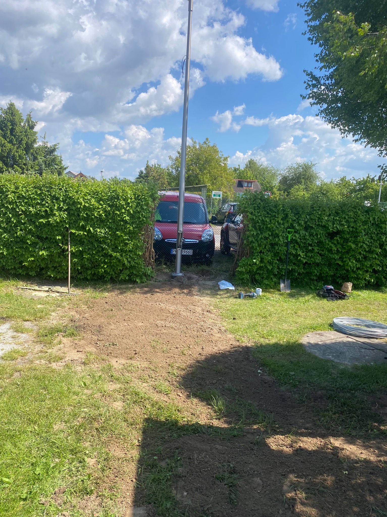 Montaż słupa oświetleniowego na posesji, widoczny czerwony samochód i narzędzia na trawie, żywopłot w tle. Słoneczny dzień.