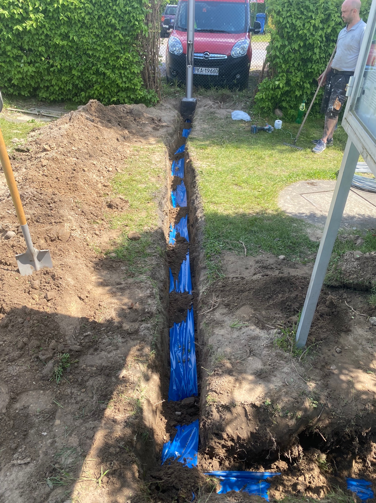 Wykop z niebieskimi rurami w ziemi, szpadel wbity w ziemię obok, trawa, krzewy i samochód w tle. Widoczny etap prac instalacyjnych.