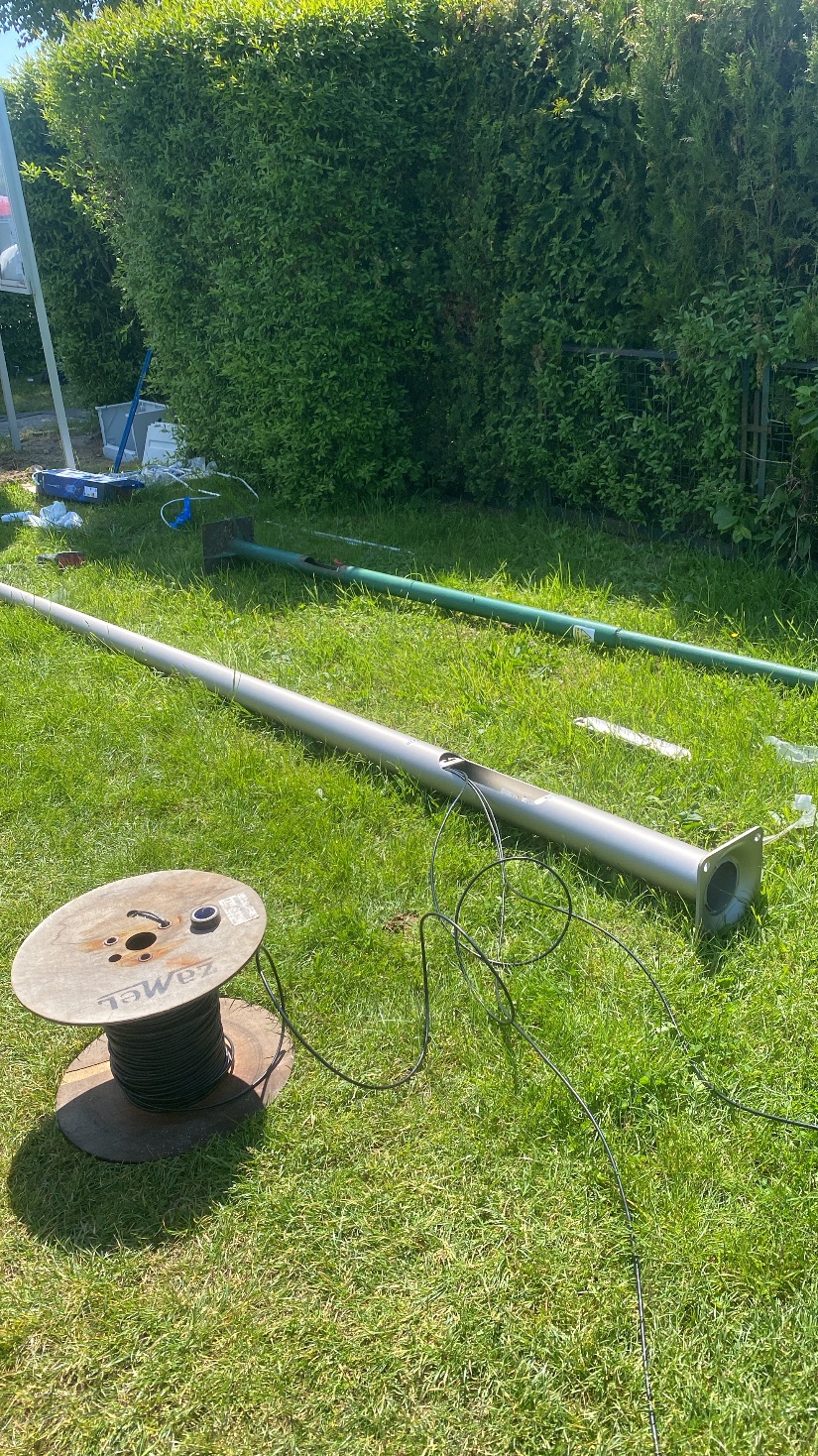 Rozłożona na trawie aluminiowa lampa ogrodowa z kablem i szpula z przewodem, przygotowywana do montażu. W tle żywopłot.