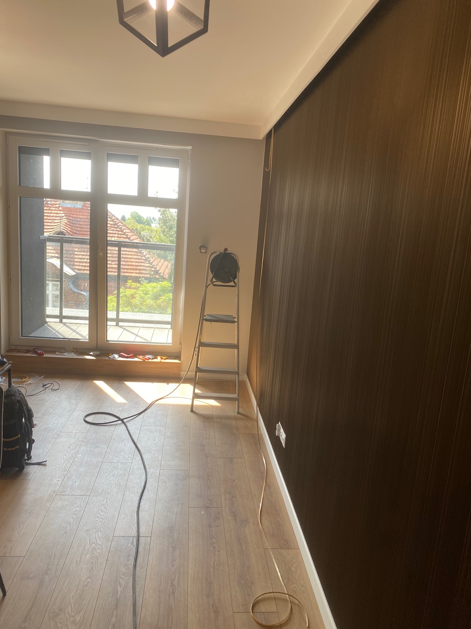 Pokój z oknem balkonowym, drabina i przewody elektryczne na podłodze. Widoczne gniazdka elektryczne na ścianie z tapetą w odcieniach brązu.