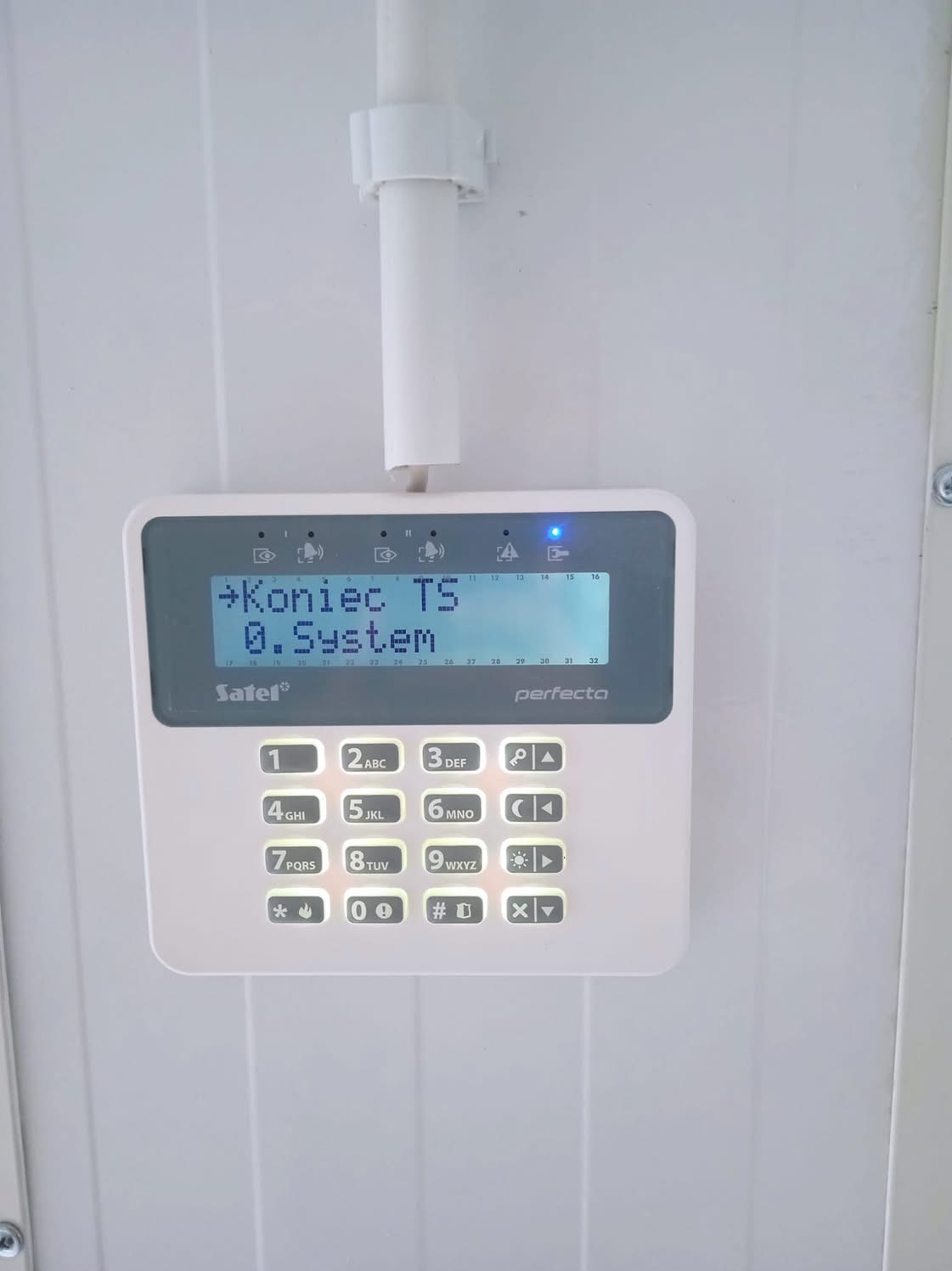 Panel sterowania alarmem Satel Perfecta z podświetlonym wyświetlaczem, zamontowany na białej ścianie z widoczną instalacją kablową.