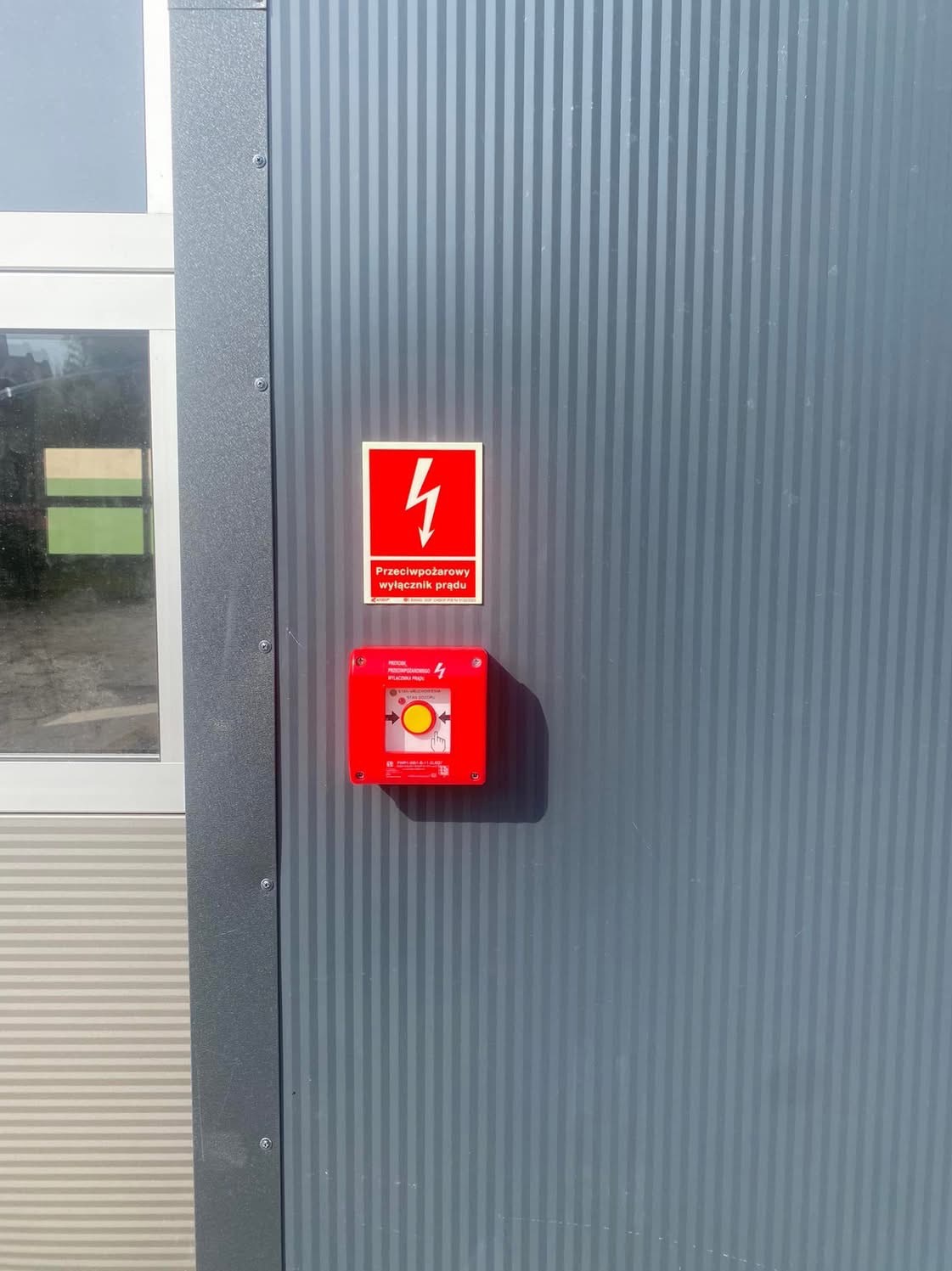 Przeciwpożarowy wyłącznik prądu z czerwoną obudową na szarej ścianie z pionowymi pasami, obok okno z szarą ramą. Oznakowanie bezpieczeństwa.