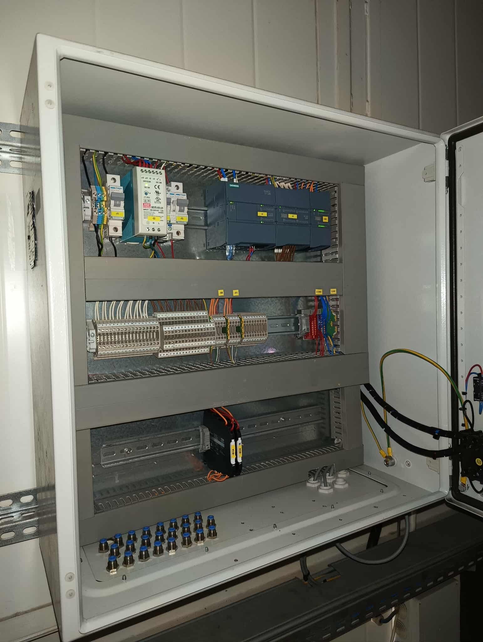 Otwarta szafa sterownicza z modułami PLC, bezpiecznikami i rozdzielnicą. Widoczne przewody, listwy zaciskowe i elementy montażowe. Szara obudowa, białe wnętrze.