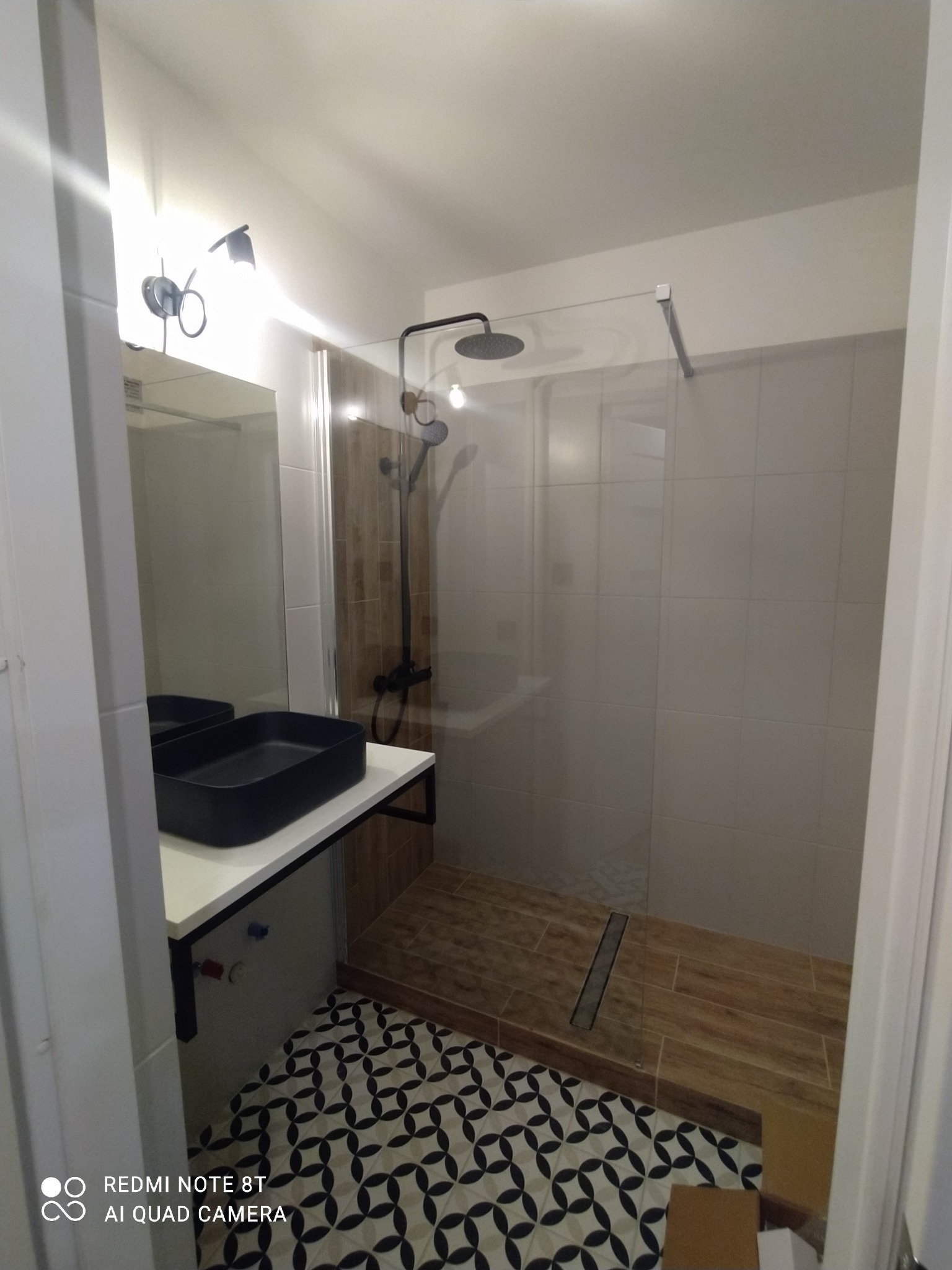 Nowoczesna łazienka z czarną umywalką nablatową, kabiną prysznicową typu walk-in z drewnopodobną podłogą i wzorzystą posadzką. Całość w jasnej tonacji.