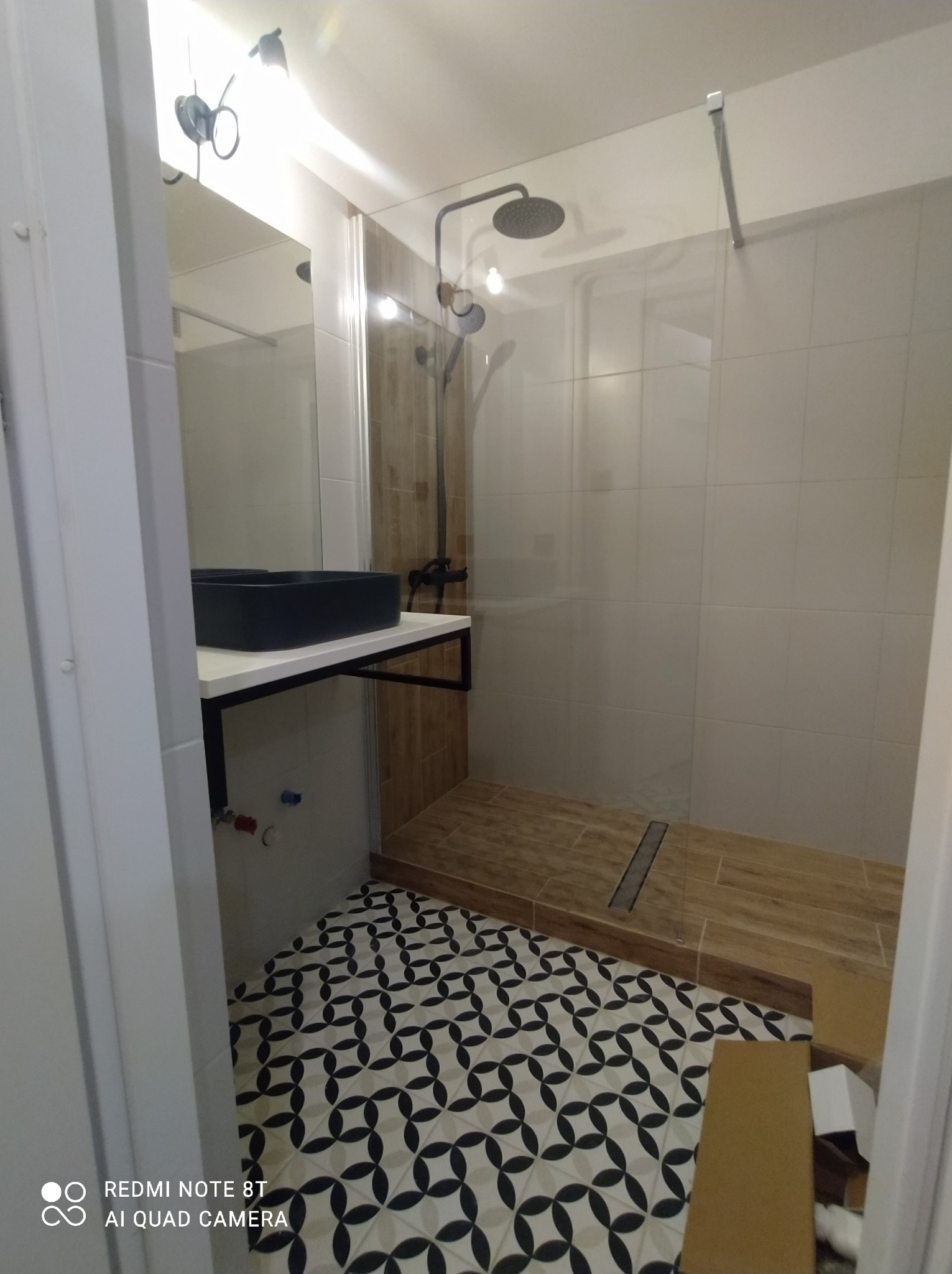 Nowoczesna łazienka po remoncie: kabina prysznicowa ze szklaną ścianą, czarna umywalka na blacie i wzorzysta podłoga. Widok zza drzwi.