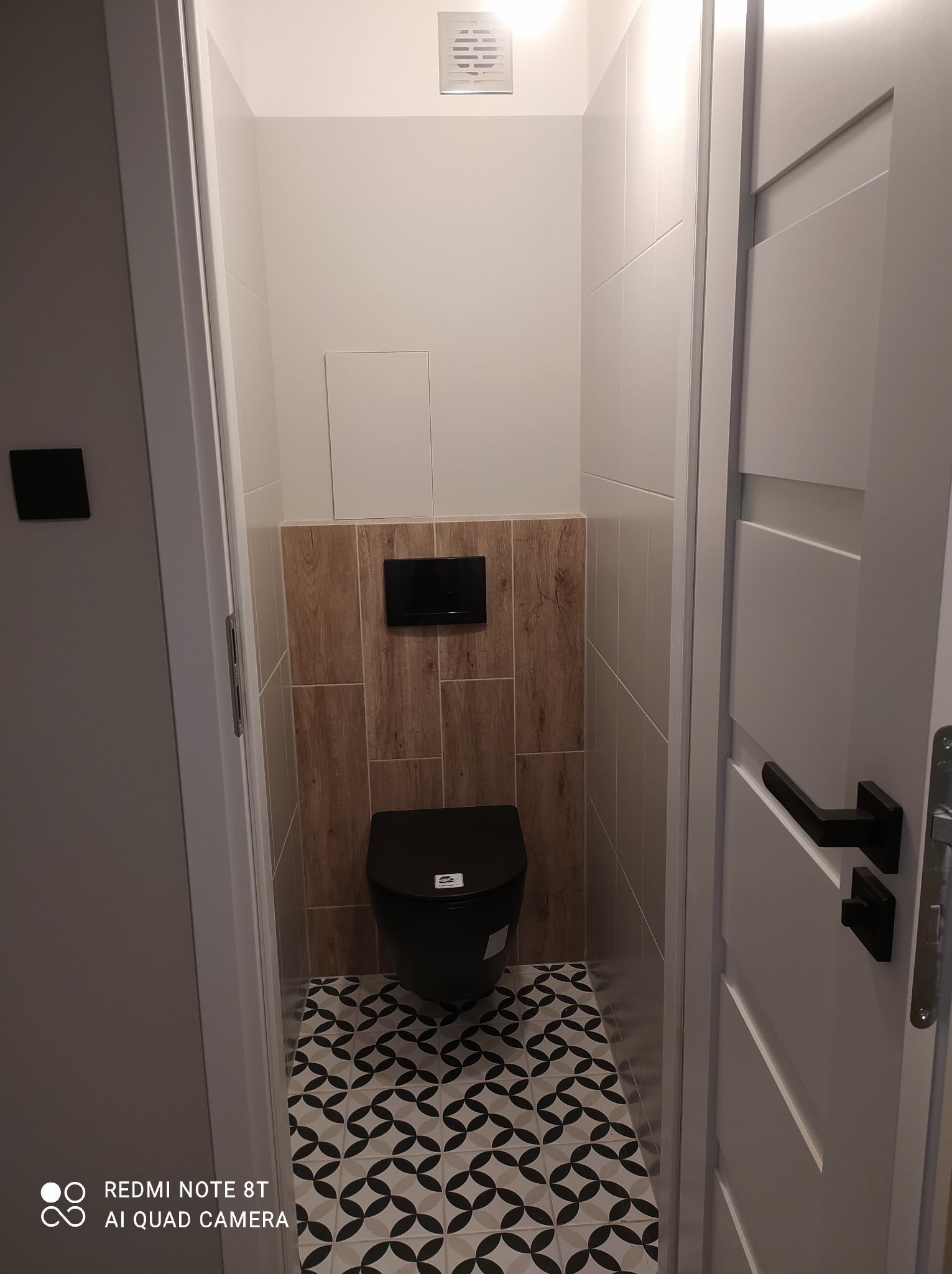 Nowoczesna toaleta w odcieniach bieli, czerni i brązu. Geometryczne płytki podłogowe, drewnopodobne panele ścienne, wisząca miska WC. Widok zza uchylonych, białych drzwi.