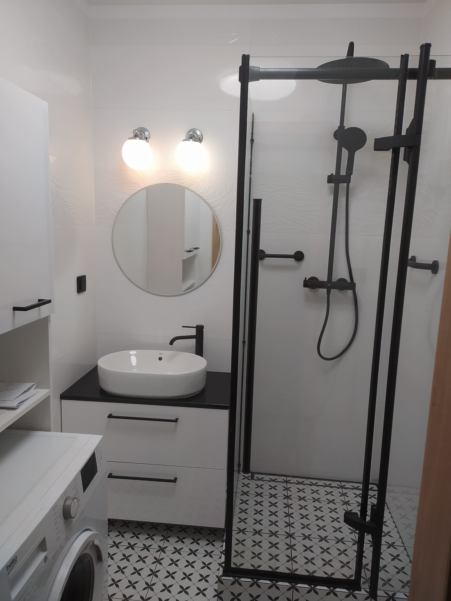Nowoczesna łazienka z białą umywalką nablatową na czarnym blacie, kabiną prysznicową z czarnymi ramami i wzorzystą podłogą. Oświetlenie punktowe i okrągłe lustro.