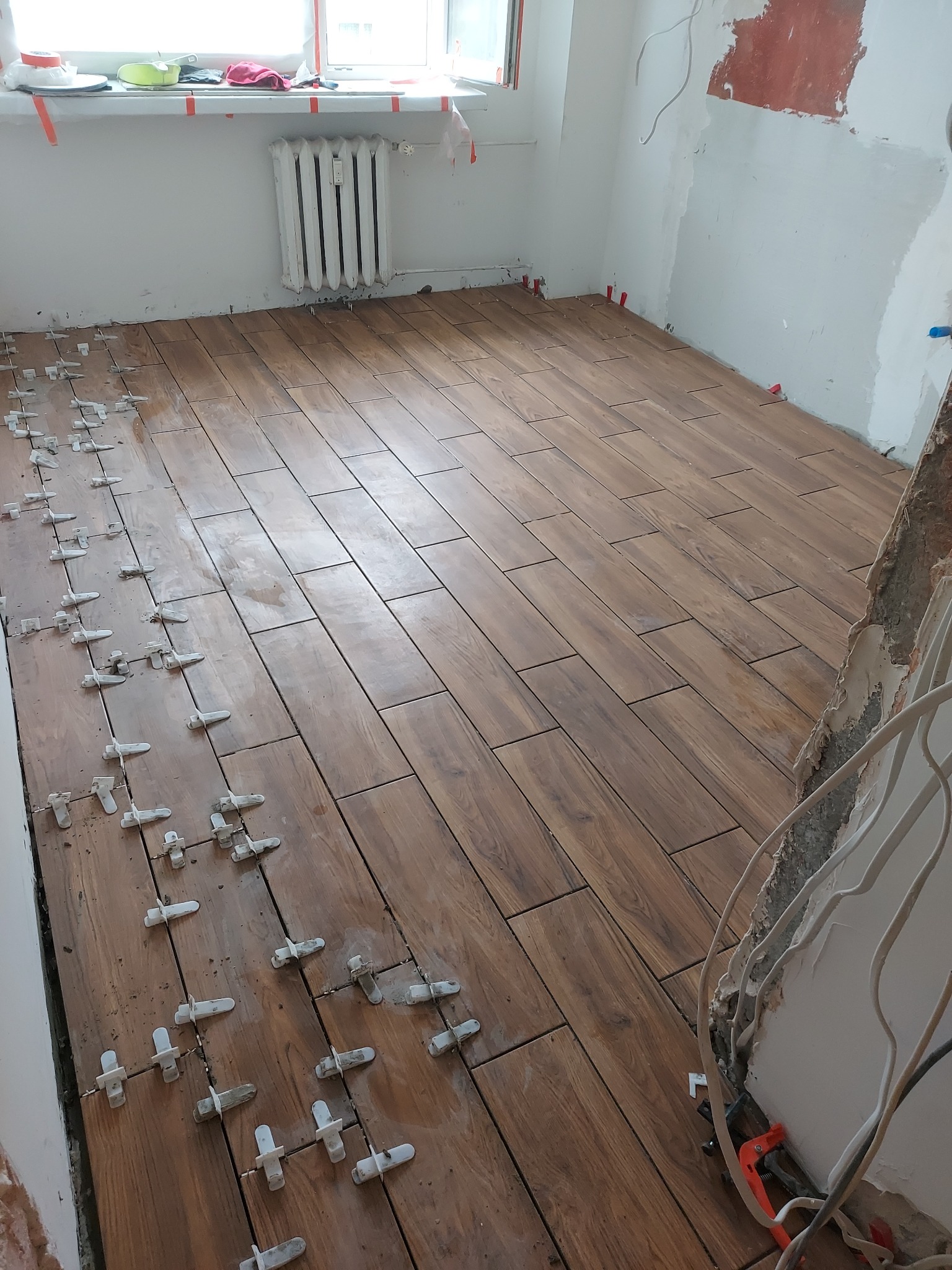 Układanie brązowych płytek imitujących drewno na podłodze w pokoju. Widoczne białe klipsy do poziomowania płytek. Częściowo wykończone ściany i okno.