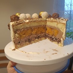 Basia Popławska - Przekrój tortu z kremem czekoladowym, mango i dekoracją z kulek kokosowych i czekoladowych, na białym talerzu obrotowym, trzymanym w dłoni.