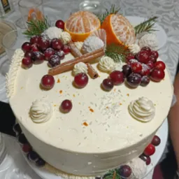 Świąteczny tort z kremem, mandarynkami, żurawiną, kokosowymi pralinkami i laskami cynamonu na białym obrusie z innymi potrawami w tle.