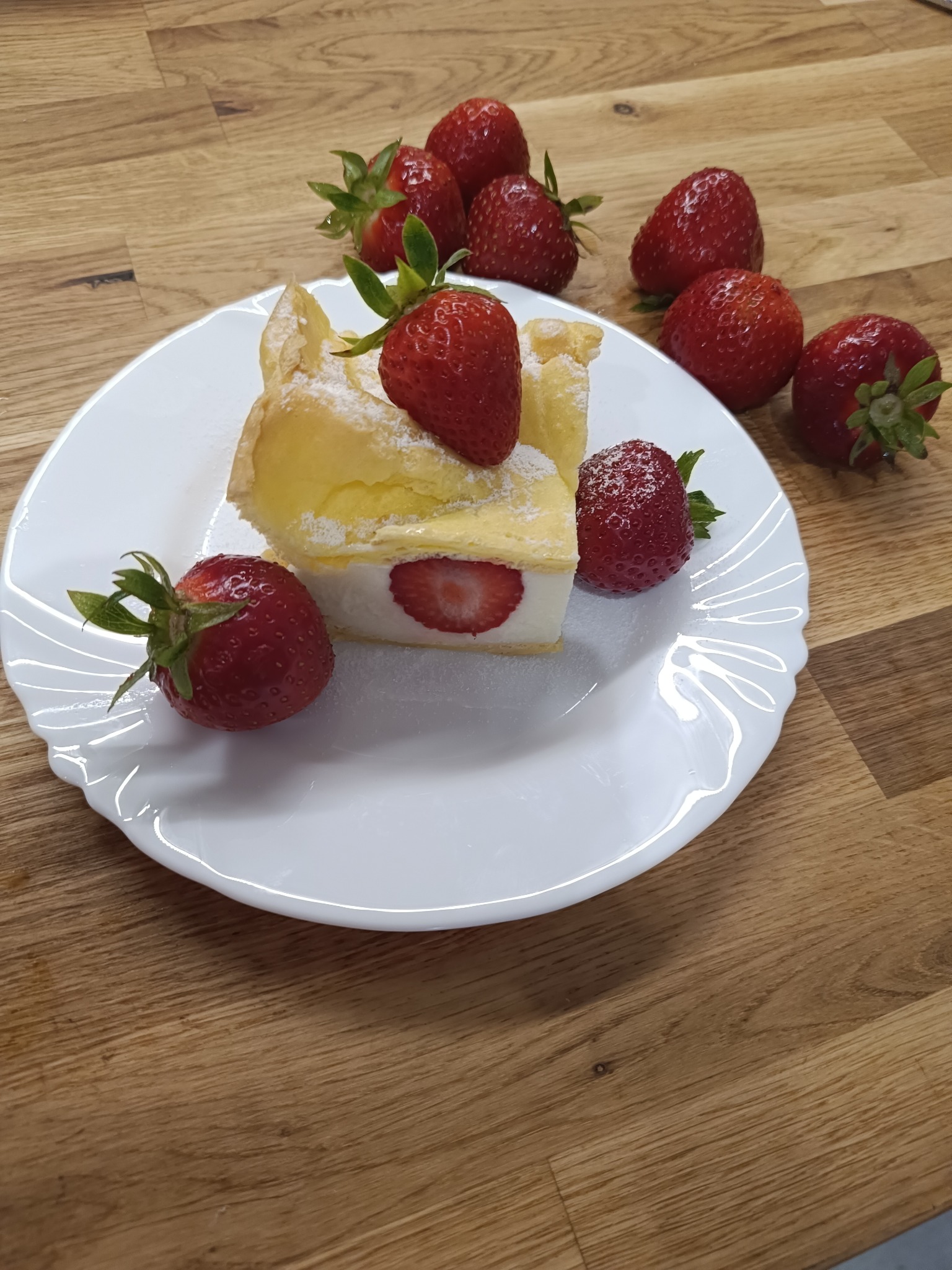 Kremówka z truskawkami na białym talerzu, posypana cukrem pudrem, na drewnianym stole. Widoczne świeże truskawki.