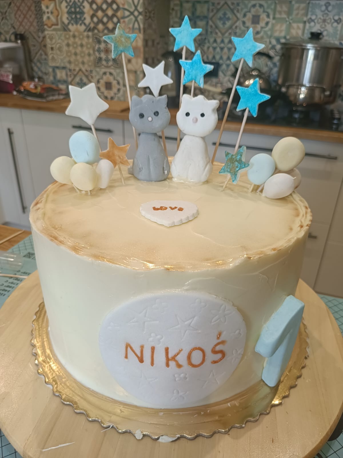 Tort urodzinowy dla Nikosia ozdobiony figurkami kotów, gwiazdkami i napisem LOVE, na drewnianym podkładzie, z elementami dekoracyjnymi w kolorach bieli, błękitu i złota.