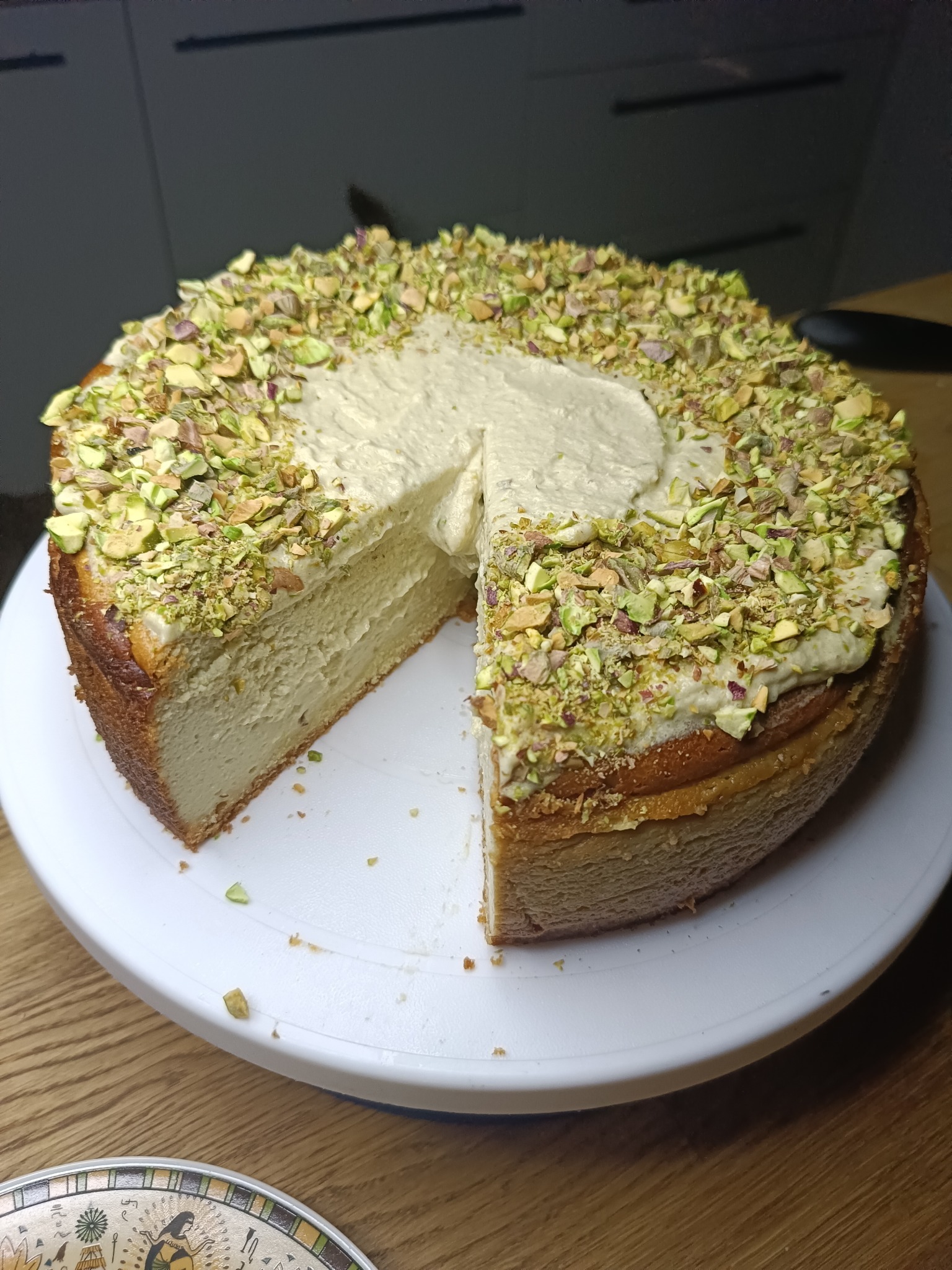 Kremowy tort z pistacjami na białym talerzu obrotowym, z wyciętym kawałkiem, ukazującym warstwy ciasta. Widoczne okruchy i drewniany stół.