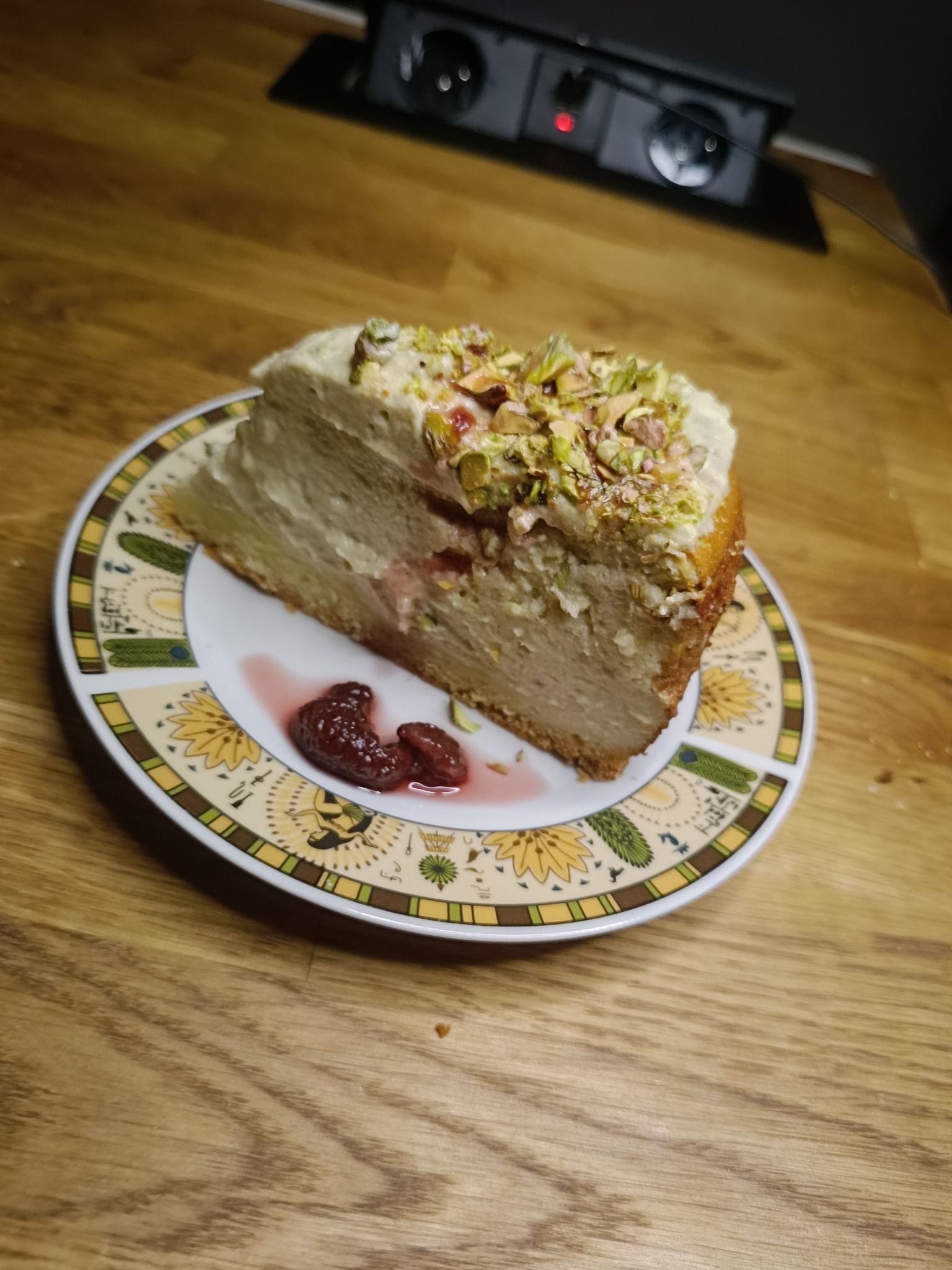 Kawałek tortu pistacjowego z kremem i dżemem na talerzu z egipskimi wzorami, na drewnianym stole. Widoczne urządzenie elektryczne w tle.