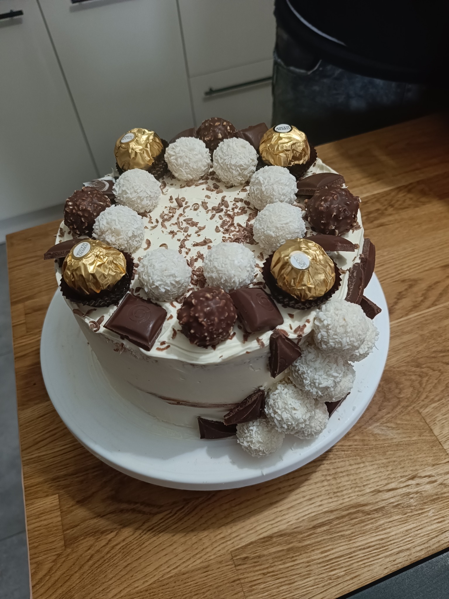 Tort z kremem, dekorowany kulkami kokosowymi, czekoladkami i pralinkami Ferrero Rocher, na drewnianym blacie. Widok z góry.