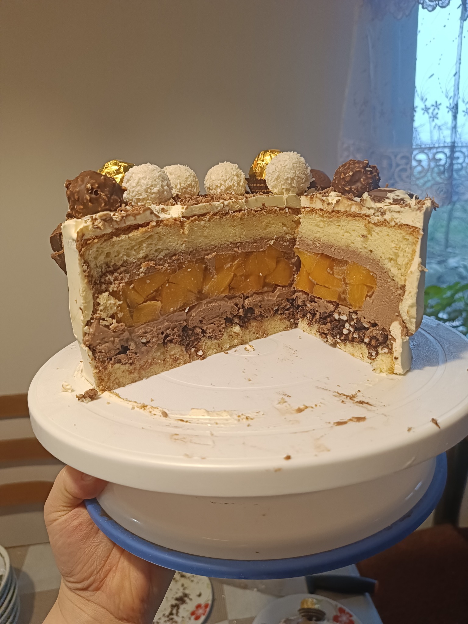 Przekrój tortu z kremem czekoladowym, mango i dekoracją z kulek kokosowych i czekoladowych, na białym talerzu obrotowym, trzymanym w dłoni.