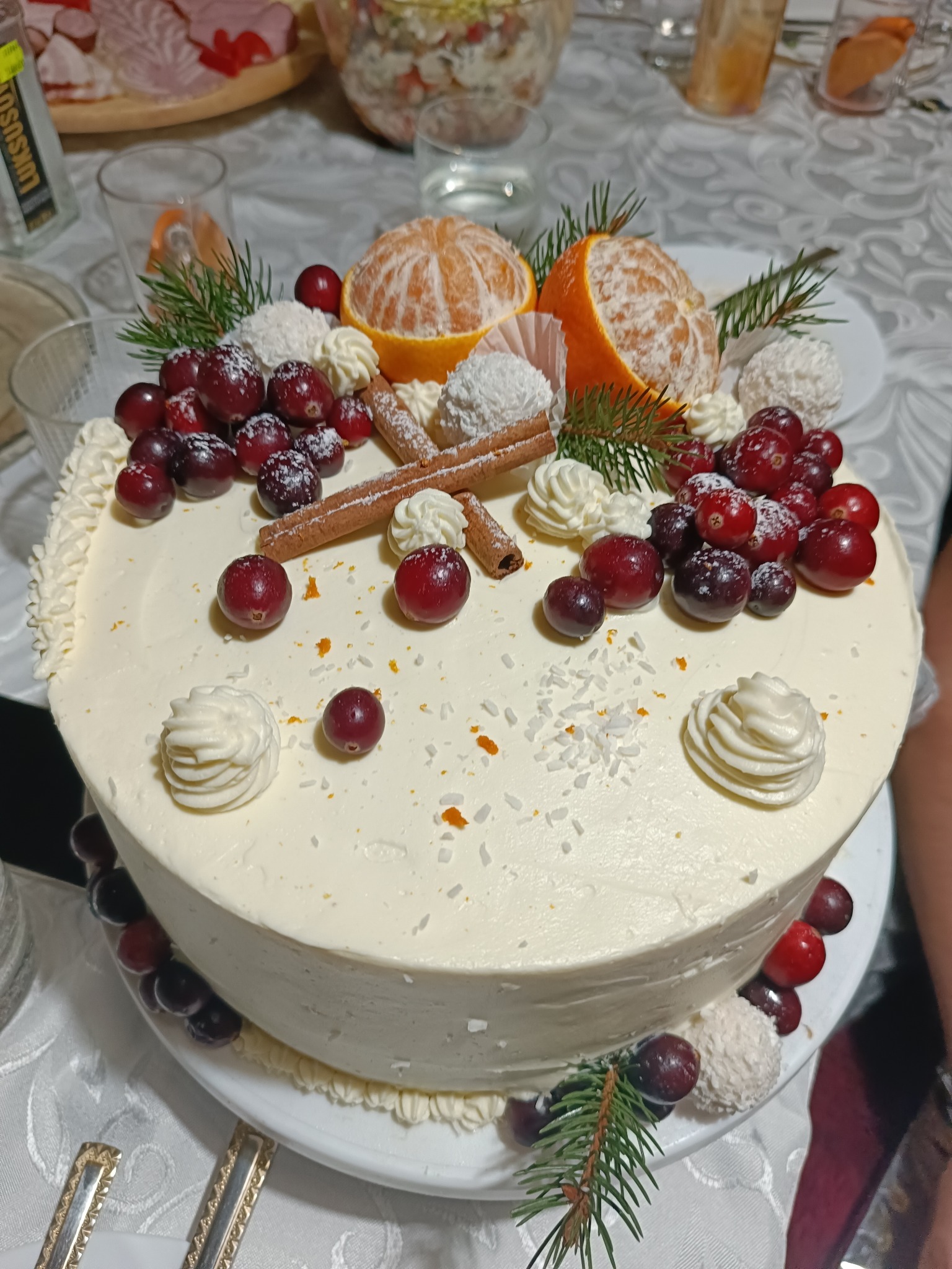 Świąteczny tort z kremem, mandarynkami, żurawiną, kokosowymi pralinkami i laskami cynamonu na białym obrusie z innymi potrawami w tle.