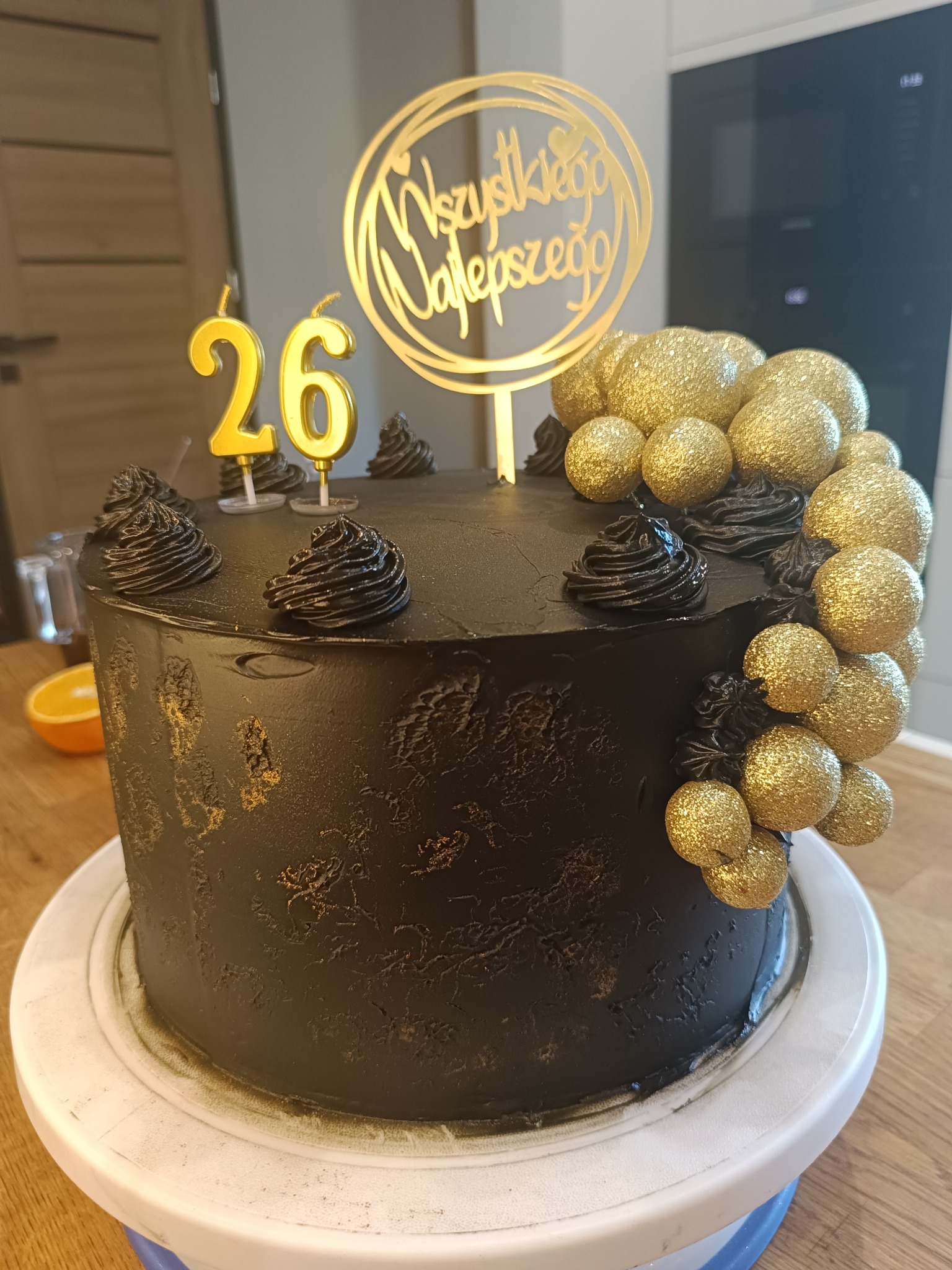 Czarny tort urodzinowy z dekoracją złotych kul, świeczkami '26' i napisem 'Wszystkiego Najlepszego' na białym stojaku, nowoczesny styl.