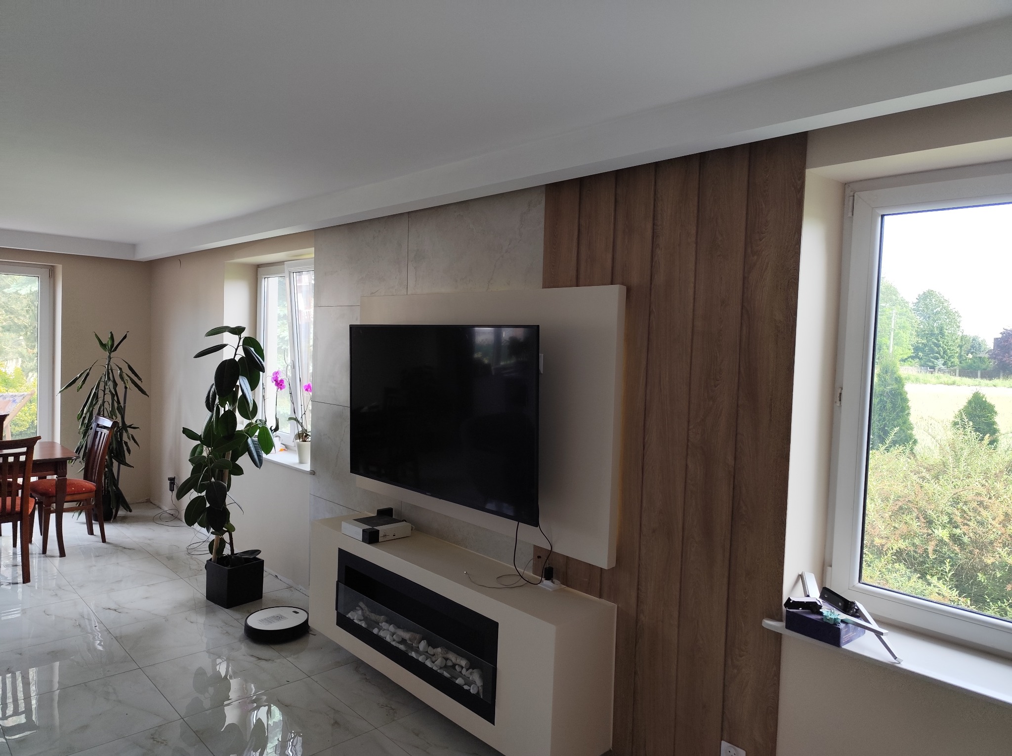 Nowoczesny salon po remoncie: ściana z panelami drewnopodobnymi i imitacją kamienia, biała szafka RTV z kominkiem elektrycznym, marmurowe płytki podłogowe.