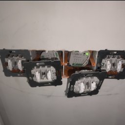 Marcin Cytarzyński Instalacje elektryczne - Gniazda i włączniki światła w trakcie montażu w ścianie, widoczne przewody i moduł sterujący F&Wave z zieloną diodą. Prace elektryczne wewnątrz budynku.