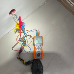 Marcin Cytarzyński Instalacje elektryczne - Pomiar instalacji elektrycznej miernikiem Sonel MPI-506. Widoczne przewody z krokodylkami podłączone do gniazdka w ścianie. Miernik stoi na podłodze obok buta.