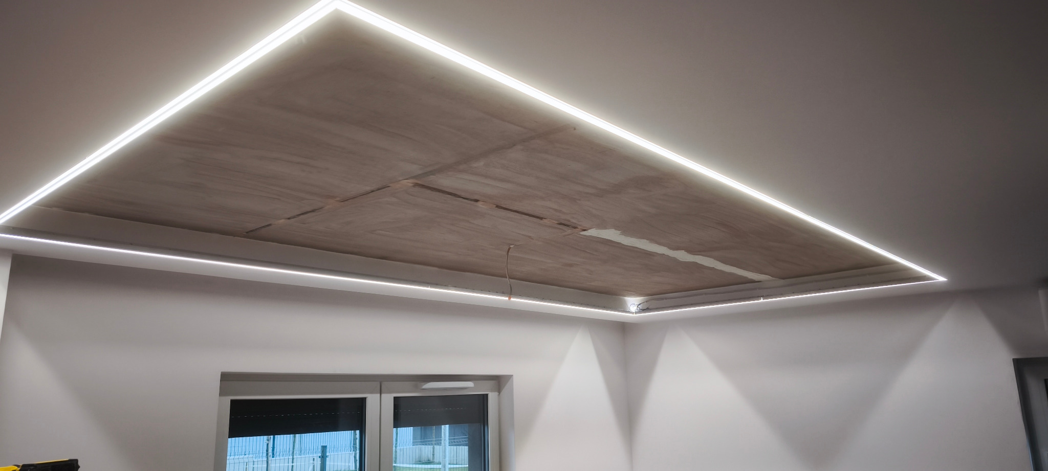 Oświetlenie LED zamontowane w suficie podwieszanym, widoczne kable. Narożnik pokoju z oknem. Surowy beton na suficie.