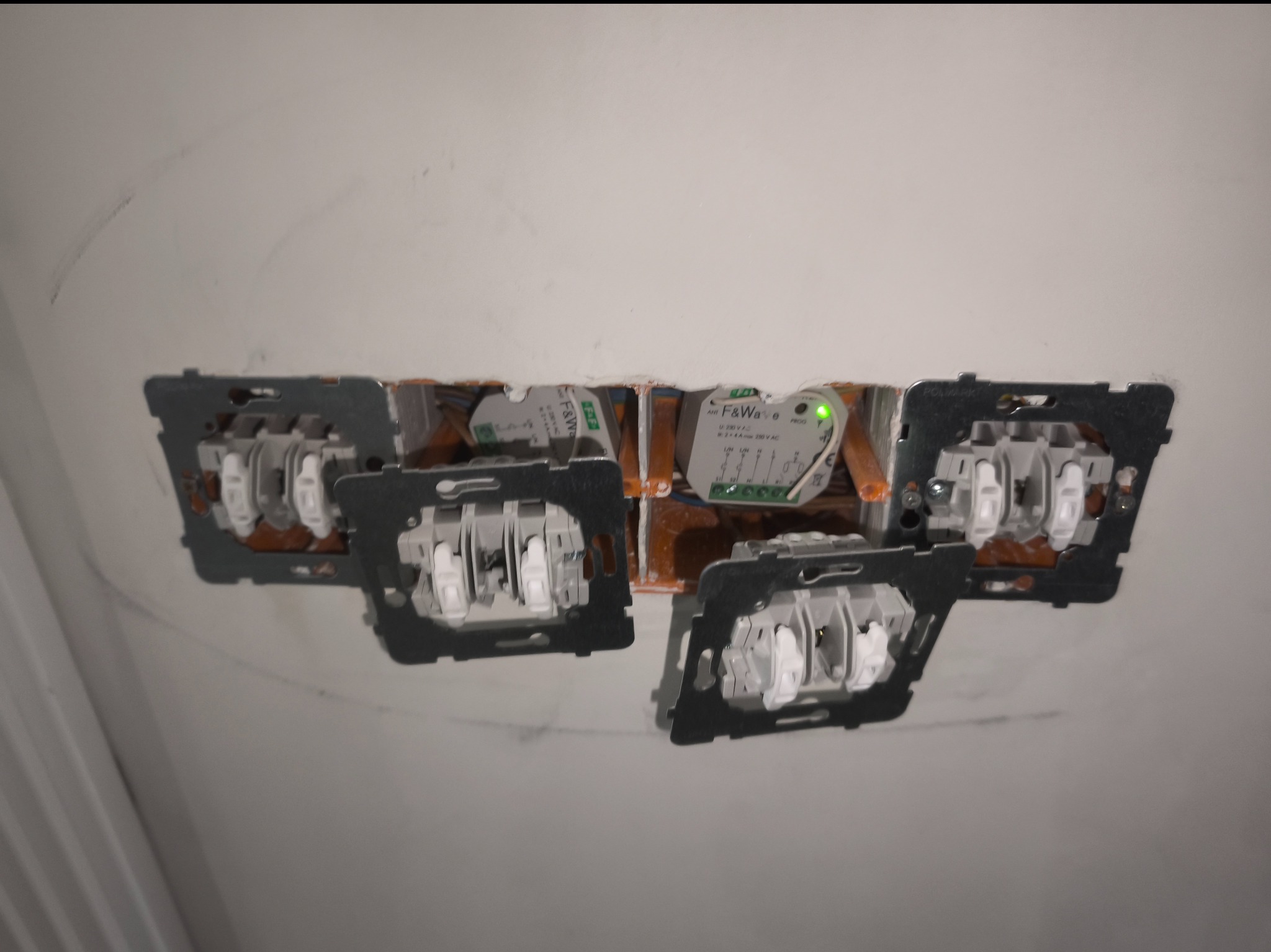 Gniazda i włączniki światła w trakcie montażu w ścianie, widoczne przewody i moduł sterujący F&Wave z zieloną diodą. Prace elektryczne wewnątrz budynku.