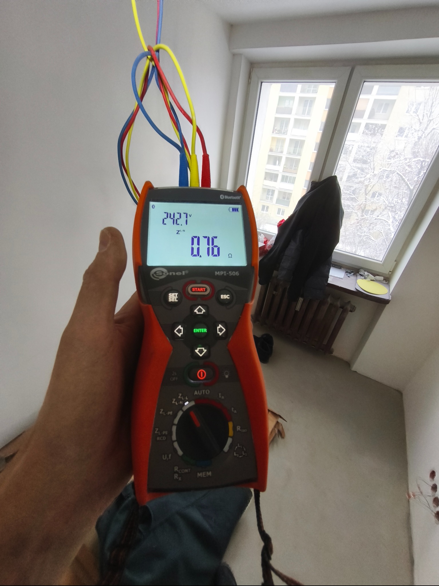 Pomiar napięcia w gniazdku elektrycznym za pomocą miernika Sonel MPI-506. Widoczne kolorowe przewody i wskazania na wyświetlaczu. W tle okno z widokiem na zimowy krajobraz.