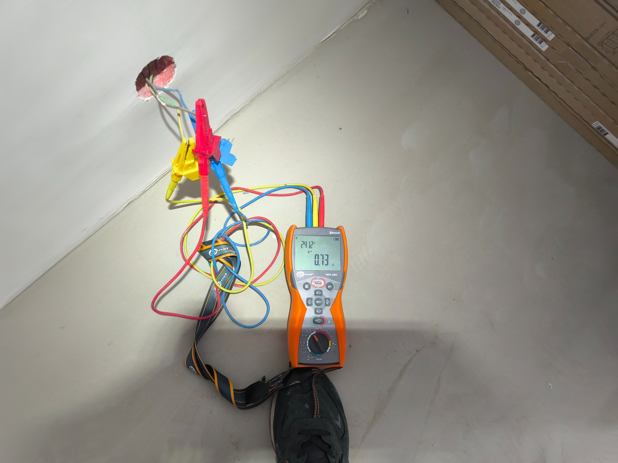 Pomiar instalacji elektrycznej miernikiem Sonel MPI-506. Widoczne przewody z krokodylkami podłączone do gniazdka w ścianie. Miernik stoi na podłodze obok buta.